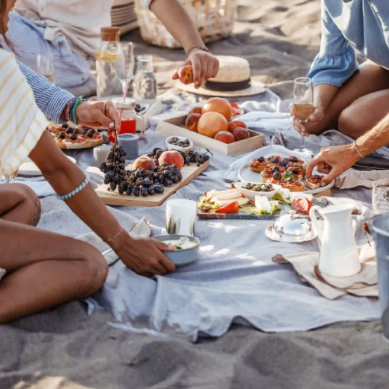 Close up shot of friends enjoying picnic on the beach with food laid out over top beach blanket 