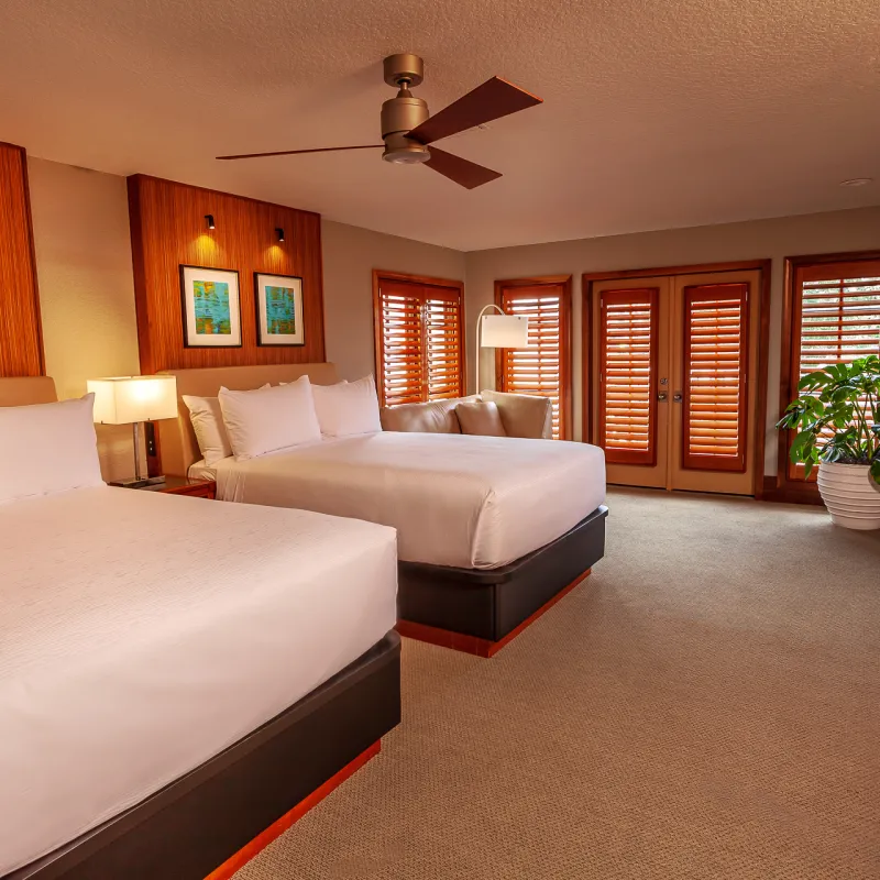 An image of a double queen bedroom in an Evermore Orlando Resort Villa. The large room features two plush queen beds, multiple lamps, a large ceiling fan, a walk mounted TV above a work desk and a set of doors that lead outside to an exterior patio.