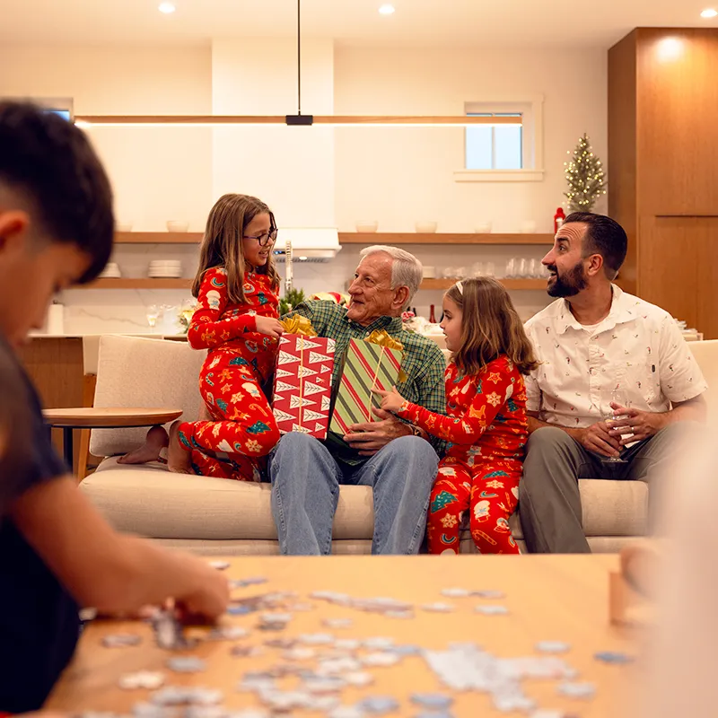 An image of kids giving dressed in Christmas pajamas giving gifts to their grandpa sitting on a couch in the living room of a house at Evermore Orlando Resort.