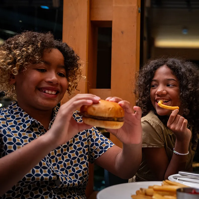 An image of two kids smiling and enjoying their food while eating dinner in Twin View Restaurant and Bar at Evermore Orlando Resort.