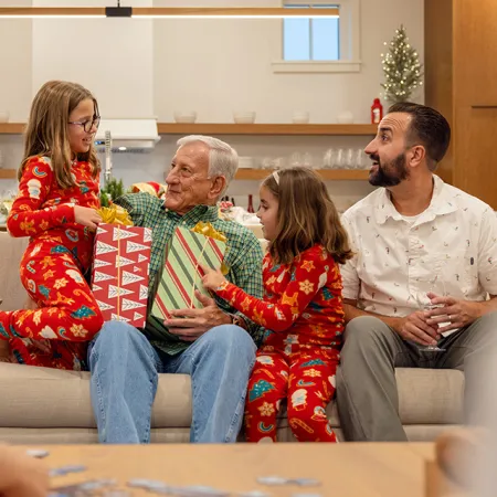 An image of kids giving dressed in Christmas pajamas giving gifts to their grandpa sitting on a couch in the living room of a house at Evermore Orlando Resort.